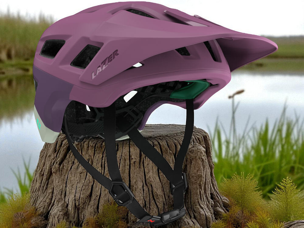 Fioletowy matowy kask rowerowy z widocznym logo \'LAZER\', umieszczony na pniu drzewa w naturalnym otoczeniu. Kask posiada otwory wentylacyjne i pasek z zapięciem.