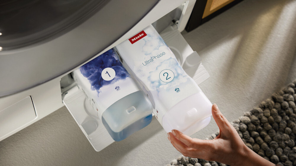Dwa pojemniki z detergentami oznaczone numerami 1 i 2 są umieszczone w szufladzie pralki. Na jednym z pojemników widoczne jest logo \'Miele\' oraz napis \'UltraPhase\'.