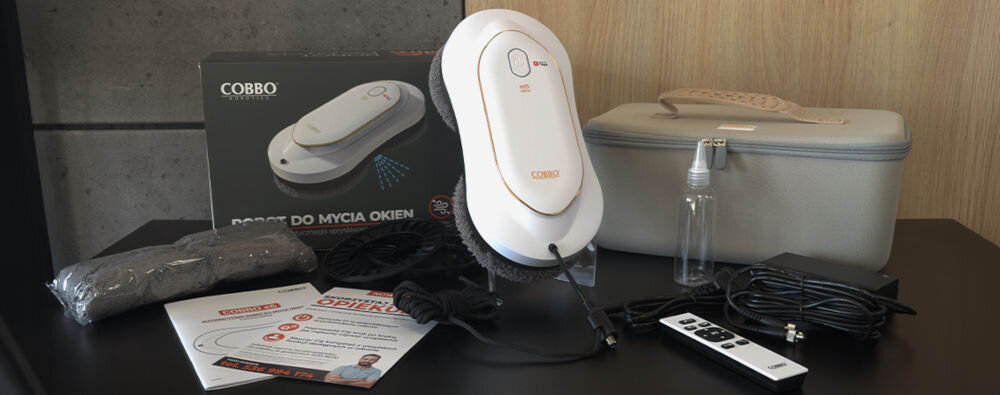 Na stole znajduje się robot do mycia okien COBBO E6 Slim wraz z akcesoriami, takimi jak pilot, przewody, butelka z rozpylaczem i instrukcje. W tle widoczny jest karton z napisem \'ROBOT DO MYCIA OKIEN\' oraz szara torba na akcesoria.