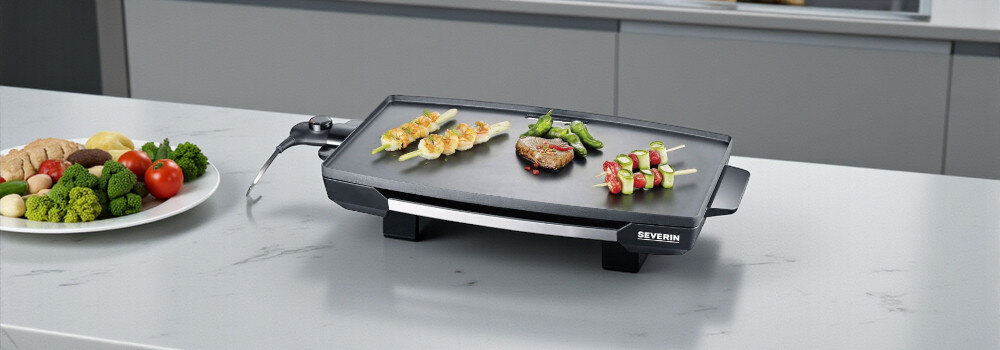 Na blacie kuchennym znajduje się grill elektryczny z logo SEVERIN, na którym grillowane są szaszłyki i kawałek mięsa. Obok grilla stoi talerz z różnymi warzywami.