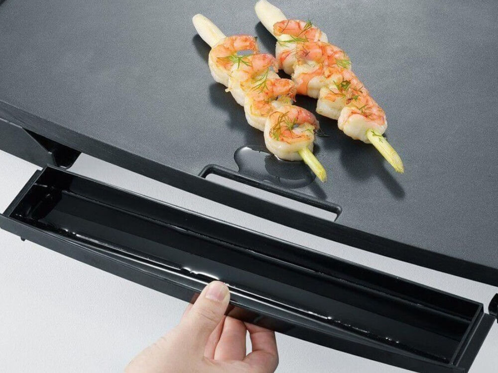 Na zdjęciu widać grill elektryczny z dwoma szaszłykami z krewetkami na powierzchni grzewczej. Osoba trzyma wysuwaną tackę na tłuszcz pod grillem.