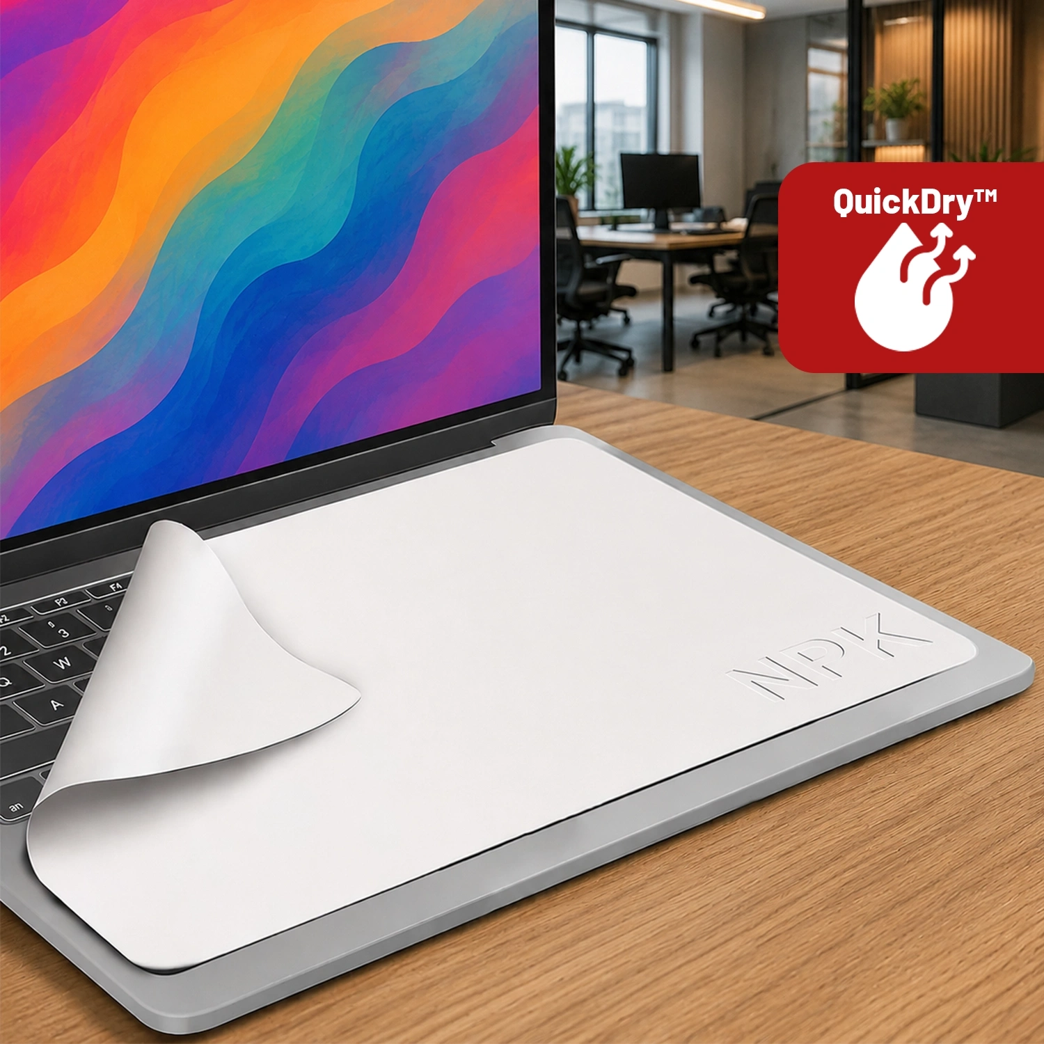 Na biurku stoi otwarty laptop z kolorowym ekranem, na klawiaturze leży biała ściereczka z napisem \'NPK\'. W prawym górnym rogu widoczna jest czerwona ikona z napisem \'QuickDry™\'.