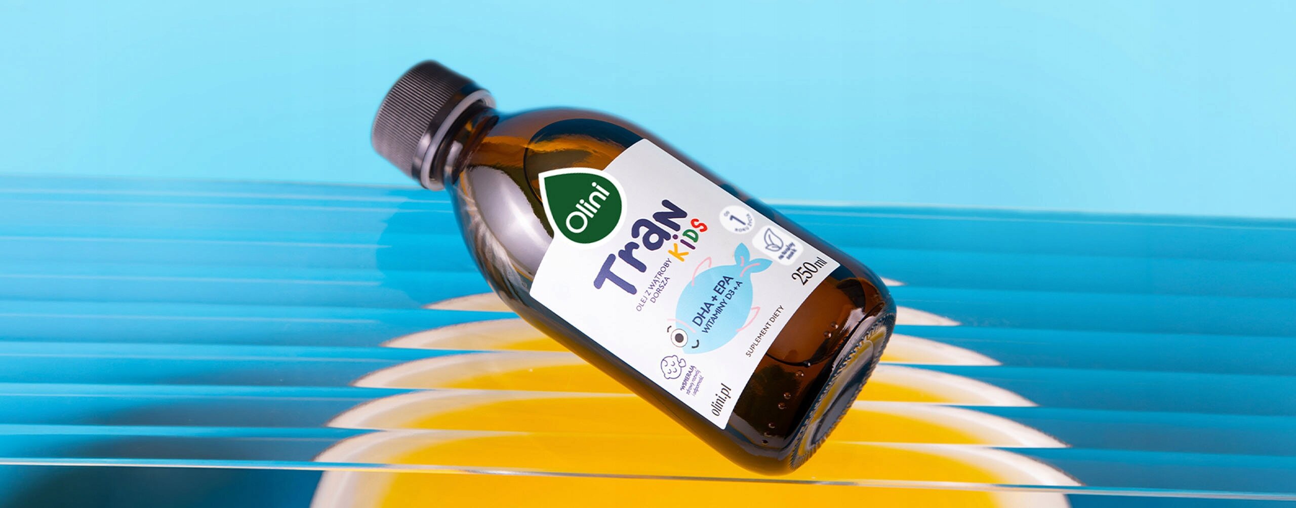 Butelka z etykietą \'Olini Tran Kids\' o pojemności 250 ml leży na kolorowym tle z niebieskimi i żółtymi odcieniami. Etykieta zawiera logo \'Olini\' oraz informacje o produkcie.