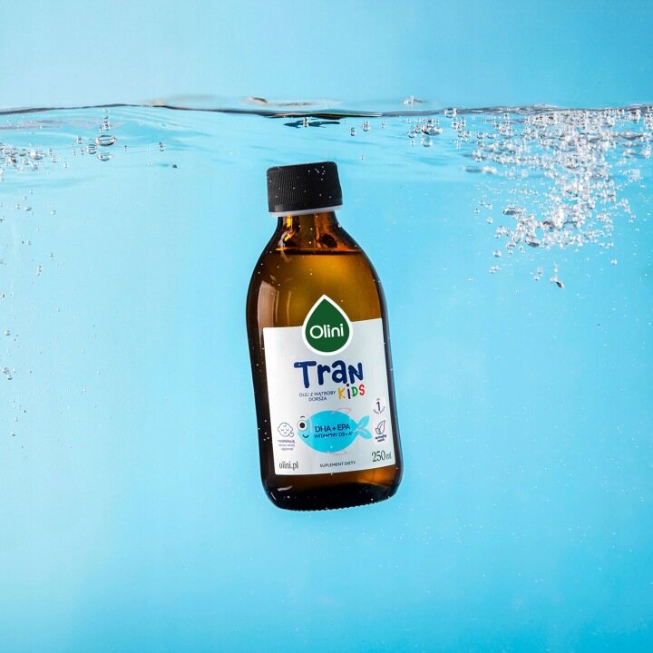 Butelka produktu Olini Tran Kids 250 ML zanurzona w wodzie, z widocznymi bąbelkami powietrza. Etykieta zawiera tekst: \'Olini Tran Kids DHA+EPA witamin D3\', a także informacje o pojemności 250 ml.