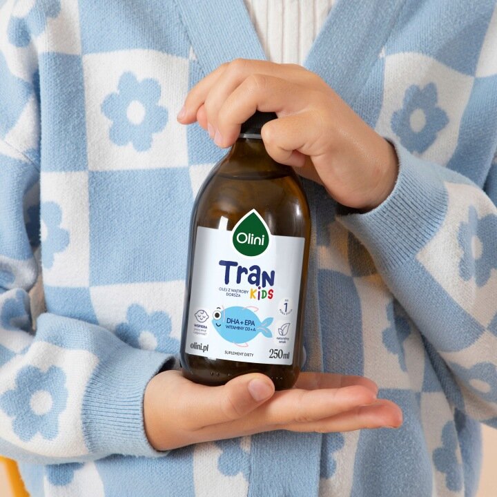 Osoba trzyma butelkę produktu Olini Tran Kids 250 ml. Etykieta zawiera informacje: \'DHA + EPA Witaminy D i A\', \'olini.pl\', \'SUPLEMENT DIETY\'.