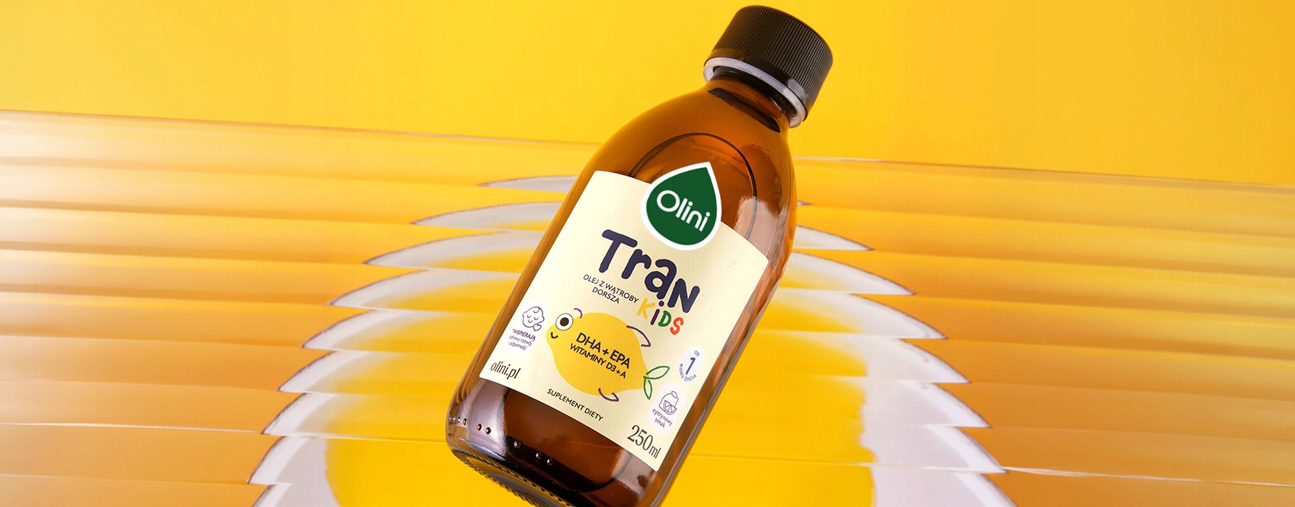 Butelka z etykietą \'Olini Tran Kids Cytrynowy 250 ML\' na tle w odcieniach żółci i bieli. Etykieta zawiera informacje o składnikach DHA i EPA oraz logo Olini.