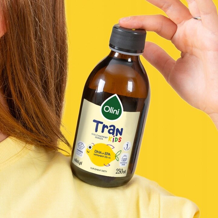 Butelka produktu Olini Tran Kids Cytrynowy 250 ml trzymana w dłoni na tle żółtej ściany. Etykieta zawiera logo Olini oraz informacje o składnikach: DHA + EPA, witaminy D3 + A.