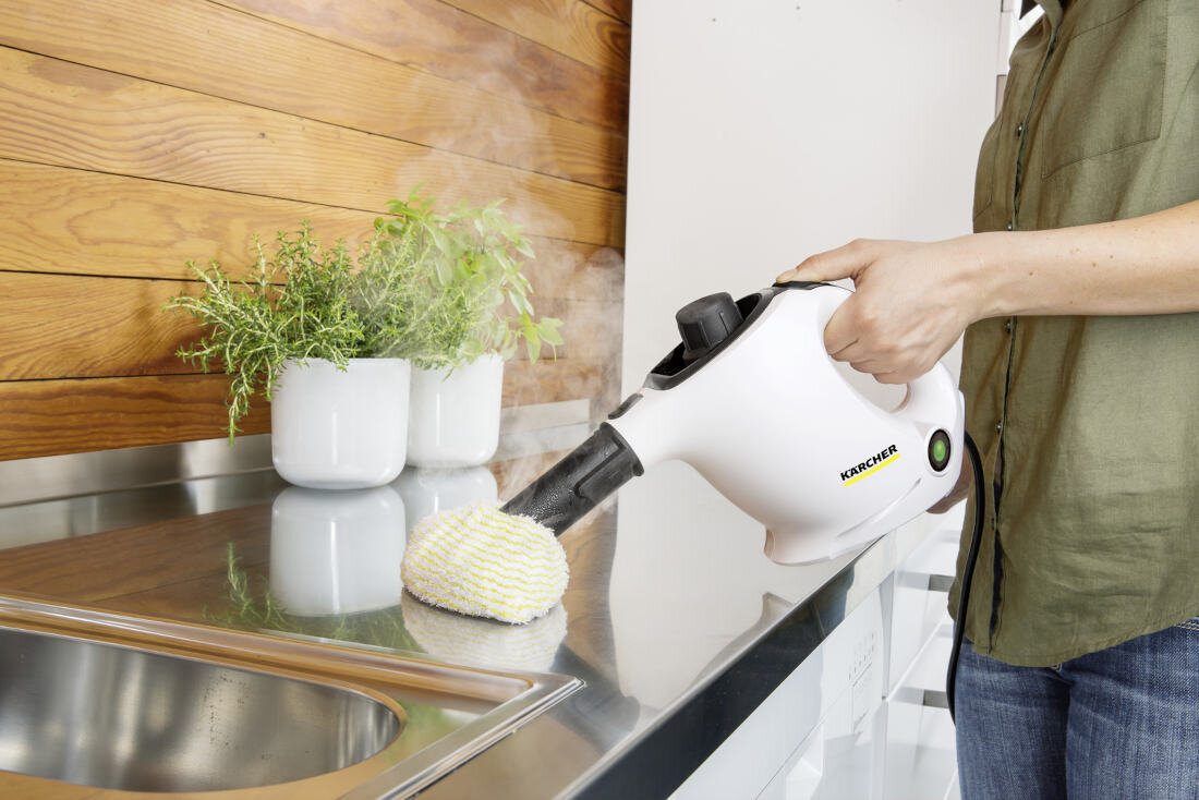 Osoba używa białej parownicy z logo \'KARCHER\' do czyszczenia metalowego blatu kuchennego. W tle widoczne są drewniane panele i białe doniczki z roślinami.