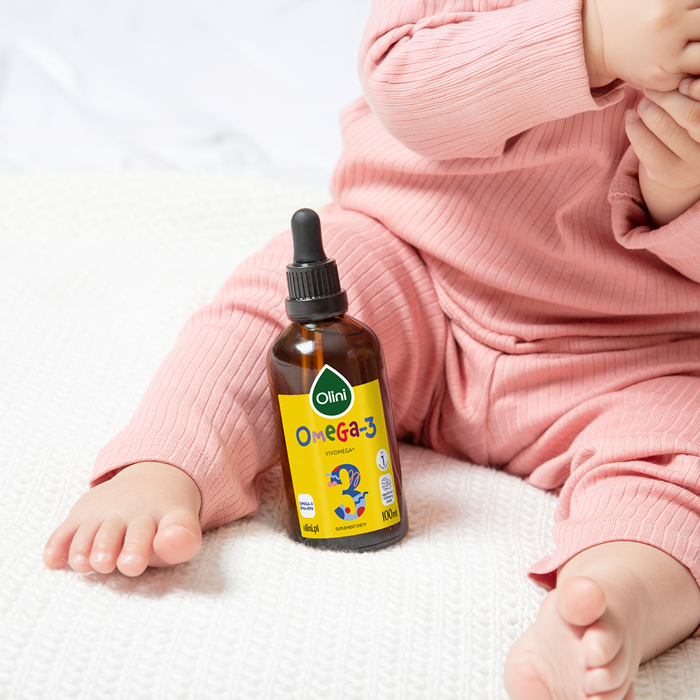 Na obrazie widoczna jest butelka z żółtą etykietą z napisem \'Olini Omega-3\' oraz dziecko w różowym ubranku siedzące na białym tle. Etykieta zawiera również grafikę rybki i napis \'100 ml\'.
