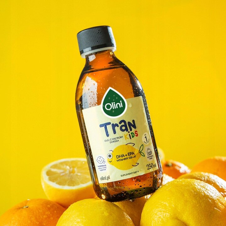 Butelka z etykietą \'Olini Tran Kids\' o pojemności 250 ml, umieszczona na tle cytryn i żółtego tła. Na etykiecie widnieje napis \'DHA+EPA Witaminy D3+K\'.