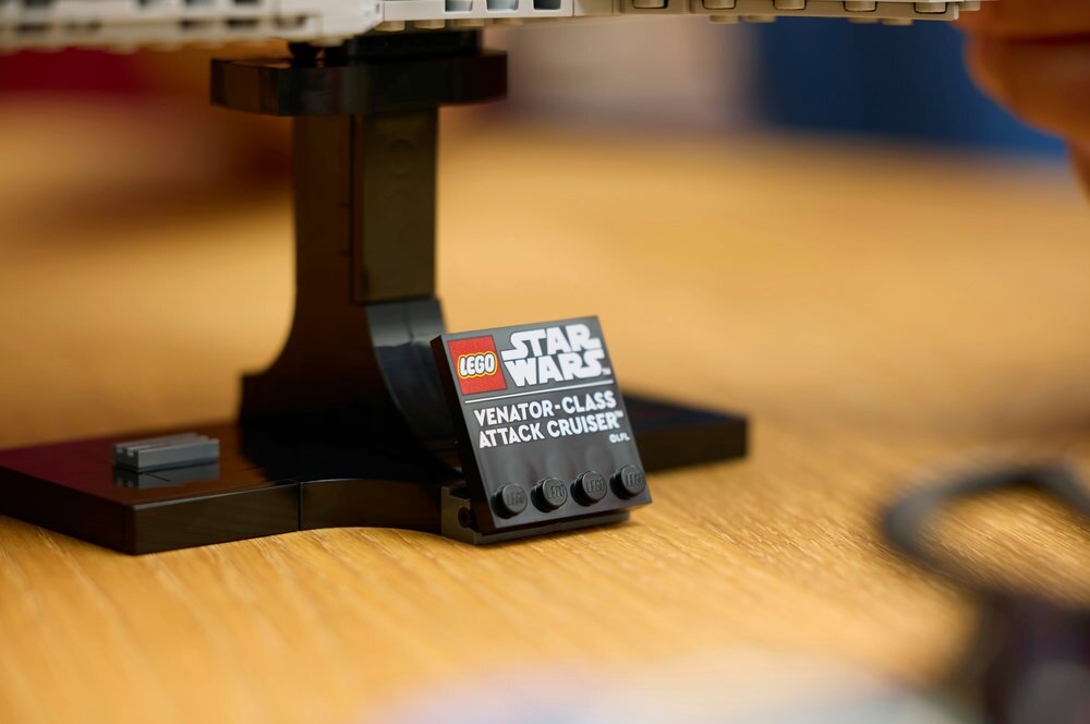 Na drewnianym blacie stoi model LEGO na czarnej podstawce z tabliczką. Tabliczka zawiera tekst: \'LEGO STAR WARS VENATOR-CLASS ATTACK CRUISER\'.