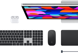 Na obrazie widoczny jest laptop z kolorowym ekranem, klawiatura, mysz oraz słuchawki bezprzewodowe. Produkty są w kolorze czarnym i srebrnym, a na myszce widoczne jest logo w kształcie nadgryzionego jabłka.