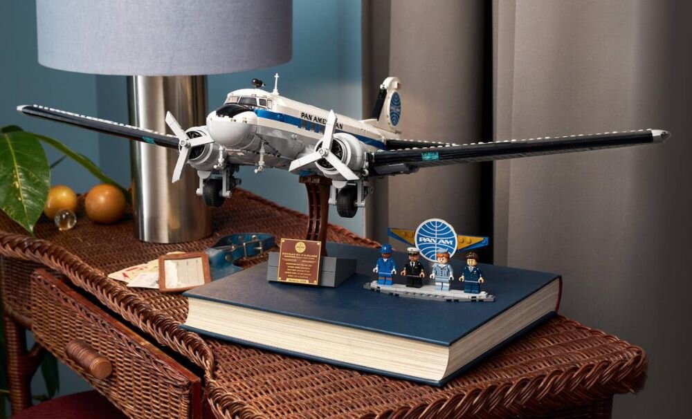 Model samolotu pasażerskiego Douglas DC-3 PAN AM z klocków LEGO ustawiony na dużej książce, obok której znajdują się figurki i tabliczka z napisem. W tle widoczna jest lampa i roślina.