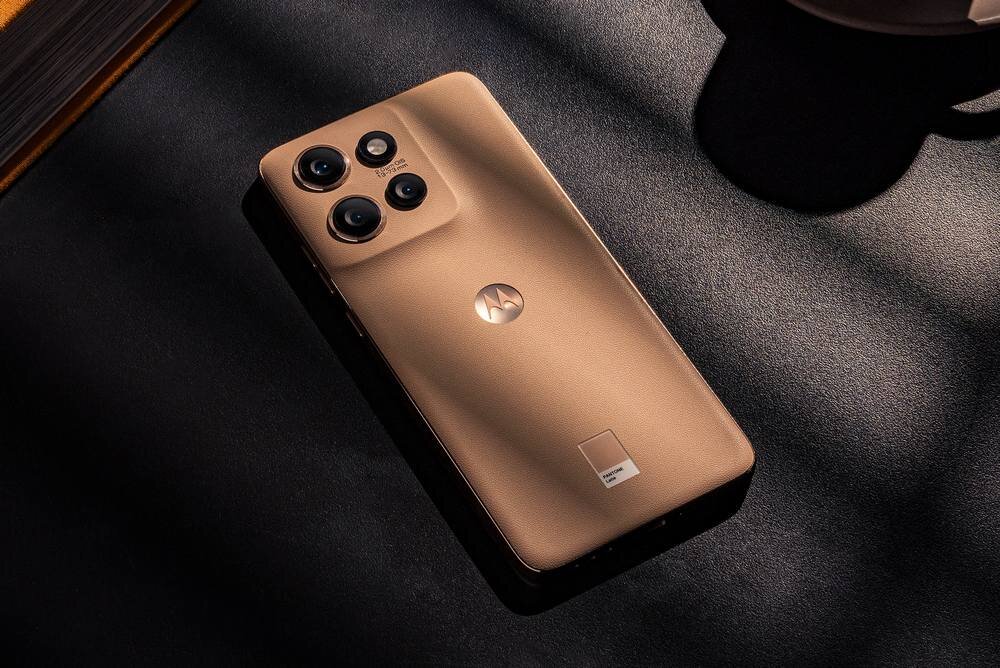 Na obrazie widoczny jest smartfon w kolorze stalowym z trzema aparatami fotograficznymi i logo producenta na obudowie. Telefon leży na ciemnej powierzchni, a światło pada na niego z góry.