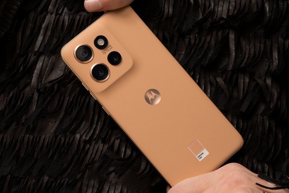 Smartfon w kolorze beżowym z trzema aparatami i logo Motorola na obudowie. Urządzenie trzymane jest w dłoni na tle ciemnego materiału.