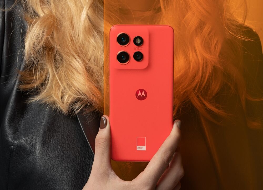 Osoba trzyma czerwony smartfon z trzema aparatami i logo Motorola na tylnej obudowie. W tle widoczne są jasne włosy i fragment czarnej kurtki.