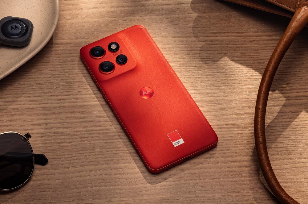 Czerwony smartfon z trzema aparatami na drewnianym stole. Obok leżą okulary przeciwsłoneczne i skórzany pasek.
