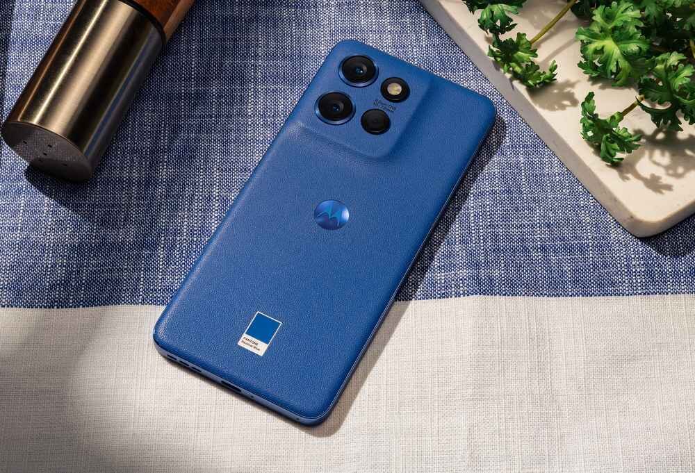 Na obrazie widać niebieski smartfon z trzema aparatami z tyłu i logo producenta. Telefon leży na tkaninie obok metalowego przedmiotu i zielonej rośliny.