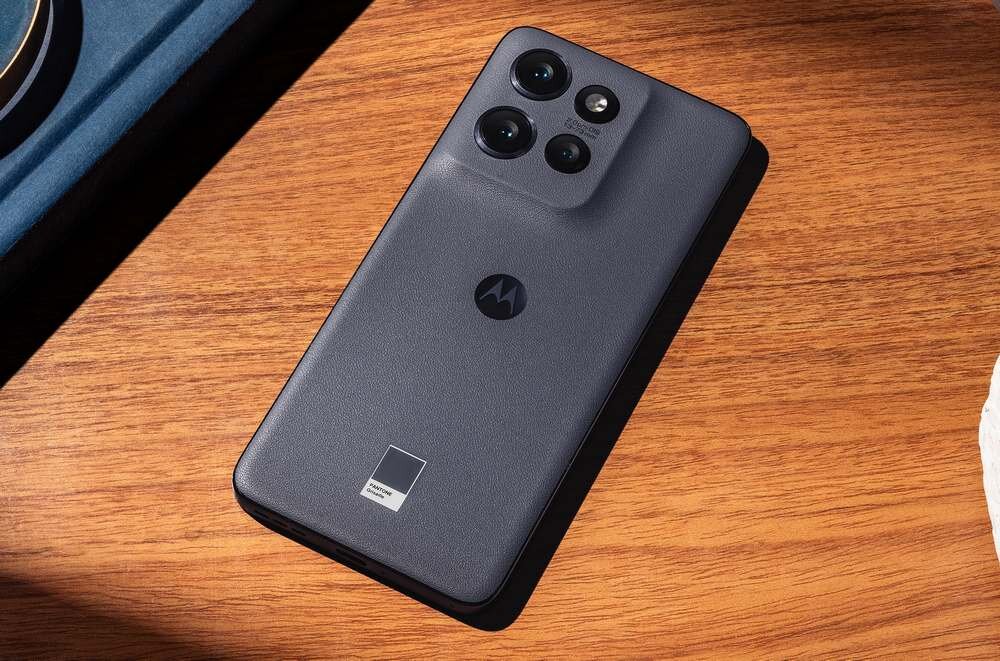 Smartfon w stalowym kolorze leży na drewnianej powierzchni, z widocznym logo producenta i trzema obiektywami aparatu z tyłu.