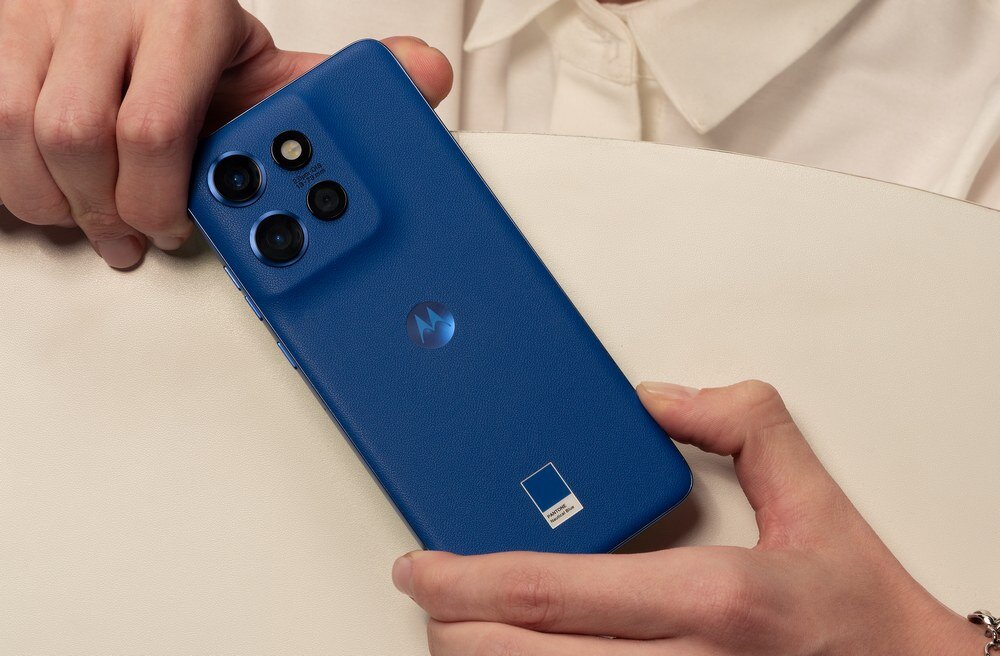 Osoba trzyma niebieski smartfon z trzema aparatami i logo Motorola na obudowie. Telefon leży na białej powierzchni.