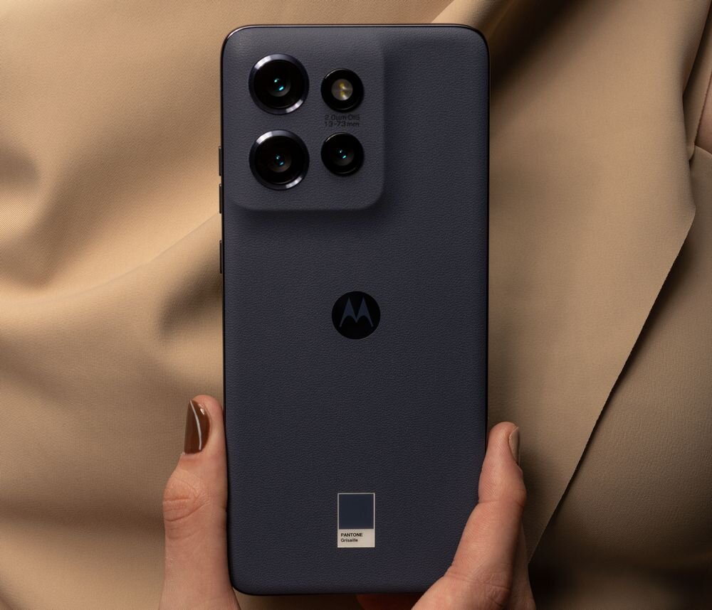 Smartfon w kolorze stalowym z trzema aparatami i logo Motorola na tylnej obudowie. W tle widoczny jest materiał w kolorze beżowym.