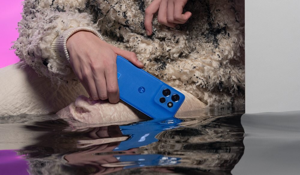 Osoba trzyma niebieski smartfon z logo w kształcie litery \'M\', częściowo zanurzony w wodzie. W tle widoczna jest różowa poświata.