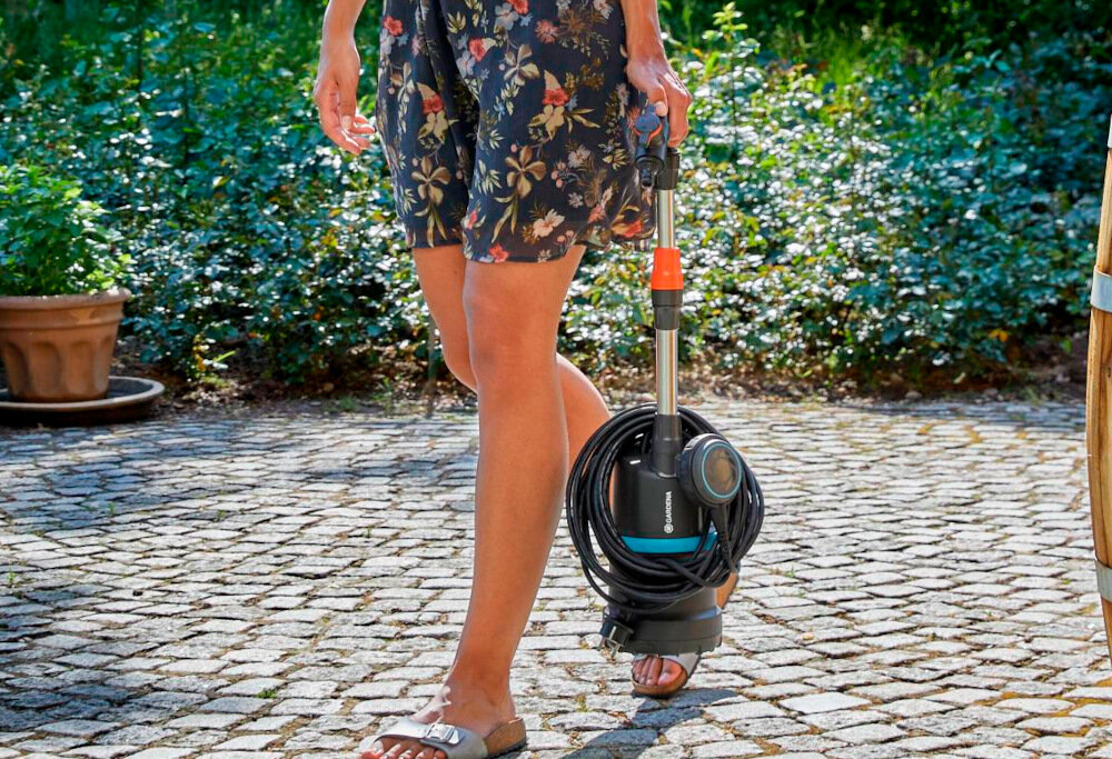 Osoba trzyma elektryczną pompę do wody marki Gardena, stojąc na brukowanej ścieżce. W tle widoczna jest roślinność i donica.