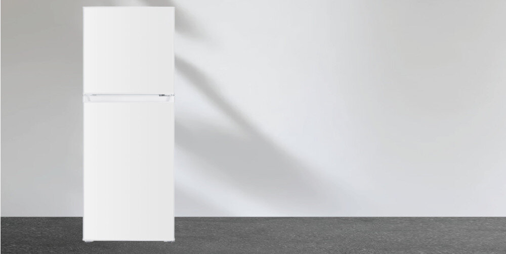 Biała lodówka z dwoma drzwiami stoi na tle jasnej ściany i ciemnej podłogi. Lodówka ma prosty, minimalistyczny design.