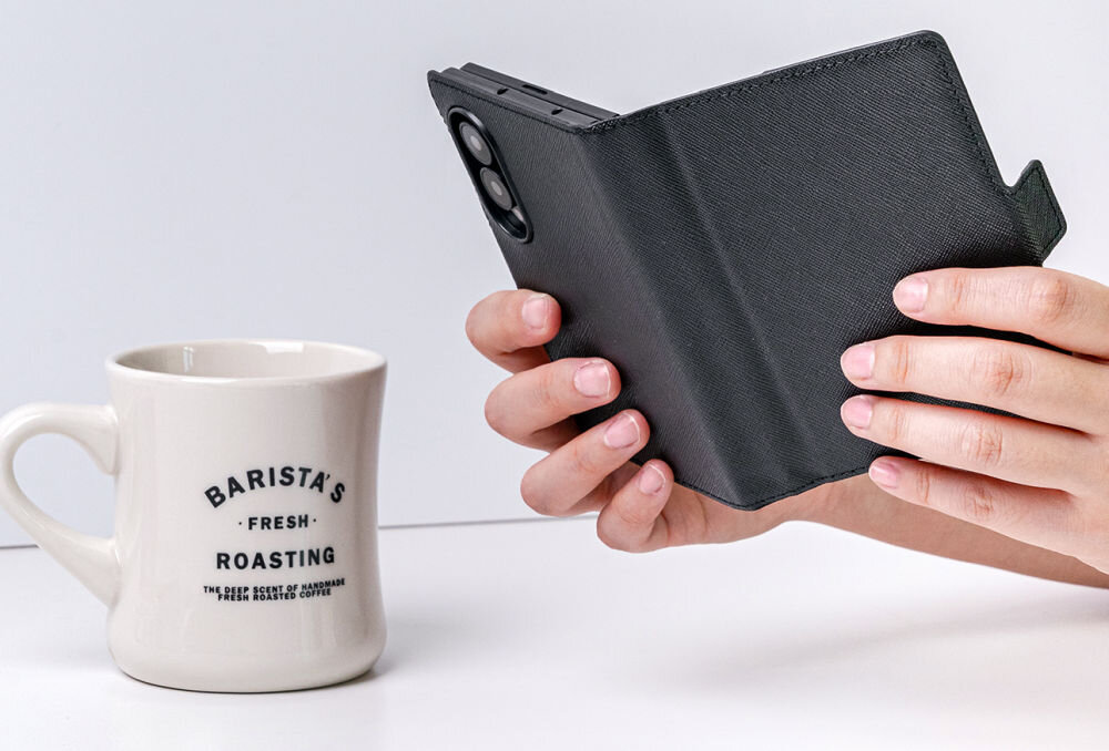 Osoba trzyma czarne etui na telefon, obok znajduje się biały kubek z napisem \'BARISTA\'S FRESH ROASTING\'.