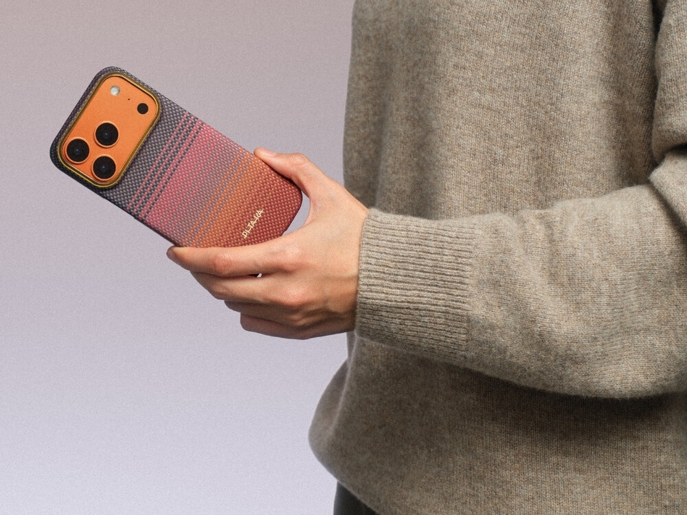 Osoba trzyma telefon w etui o gradientowym wzorze w odcieniach pomarańczu i różu. Na etui widoczny jest napis \'PITAKA\'.