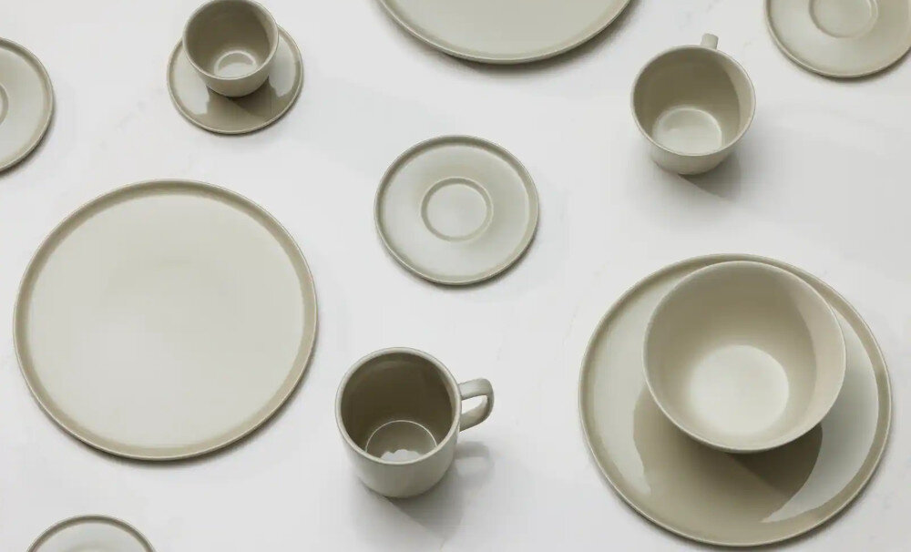 Na obrazie widoczny jest zestaw porcelanowych naczyń w jasnym kolorze, w tym talerze, filiżanki i spodki, ułożone na białym tle.