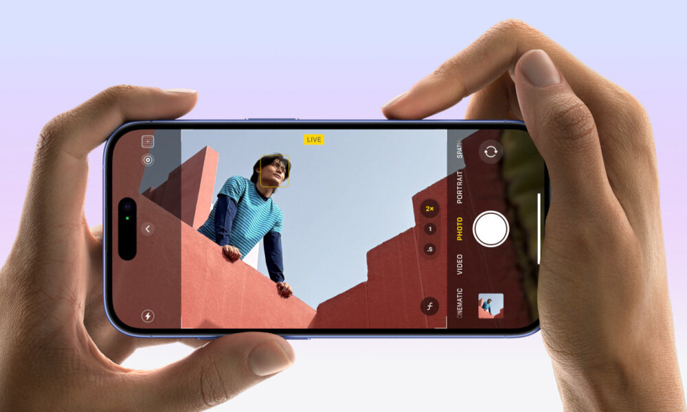 Osoba trzyma smartfon, na ekranie którego widać aplikację aparatu z podglądem zdjęcia przedstawiającego osobę na tle czerwonej ściany. Na ekranie widoczne są ikony trybów aparatu oraz przycisk migawki.