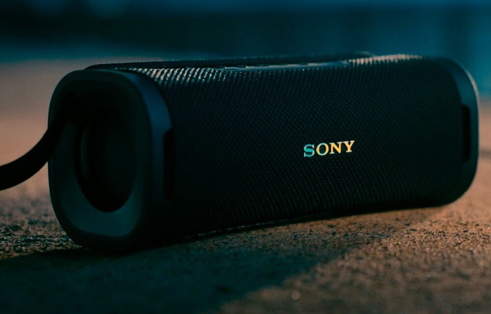 Czarny głośnik mobilny z widocznym logo \'SONY\' na siatkowanej powierzchni. Urządzenie leży na płaskiej powierzchni w przyciemnionym oświetleniu.