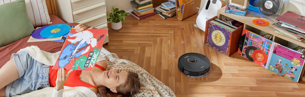 Na drewnianej podłodze znajduje się czarny robot sprzątający. W tle widać półki z kolorowymi płytami winylowymi i osobę leżącą na dywanie, trzymającą jedną z płyt.