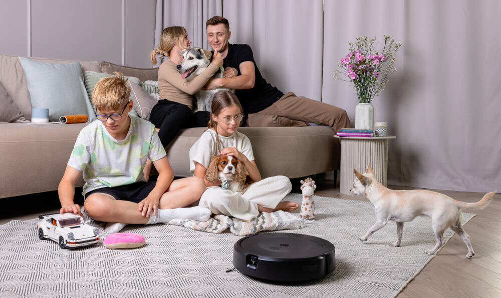 Na dywanie znajduje się robot sprzątający. W tle widoczna jest kanapa i stolik z wazonem pełnym kwiatów.