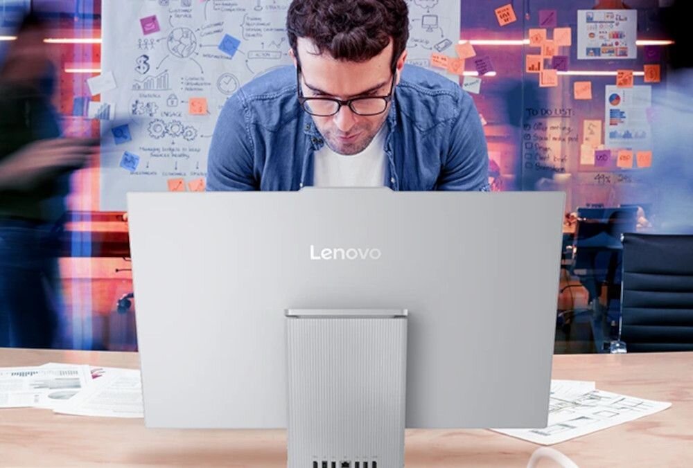 Mężczyzna pochyla się nad biurkiem, na którym stoi komputer All in One z widocznym logo Lenovo. W tle znajdują się tablice z notatkami i rysunkami.