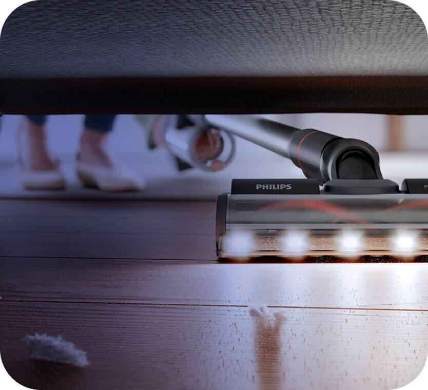 Odkurzacz pionowy z widocznym logo PHILIPS, oświetlający podłogę pod meblem. W tle widać rozmytą postać w butach.