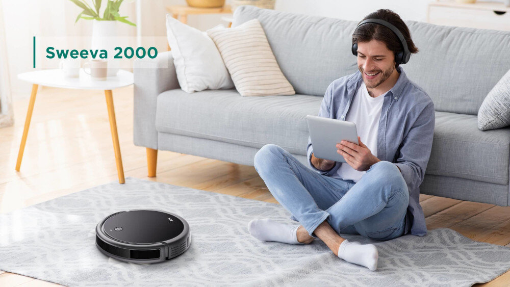 Na dywanie w salonie znajduje się czarny robot sprzątający. Obok siedzi osoba z tabletem, a w tle widać kanapę i stolik z rośliną.