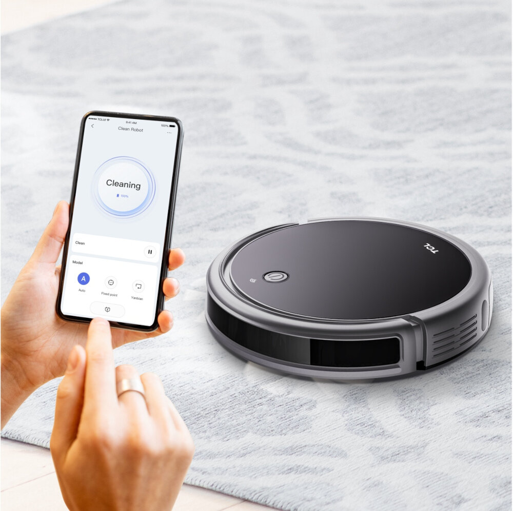 Na dywanie znajduje się czarny robot sprzątający z logo TCL. Obok osoba trzyma smartfon z aplikacją pokazującą status \'Cleaning\'.