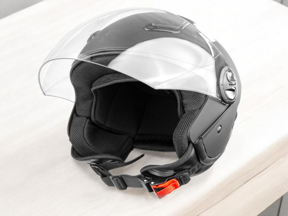 Czarny kask motocyklowy z przezroczystą szybką i czerwonym zapięciem, leży na jasnym stole.