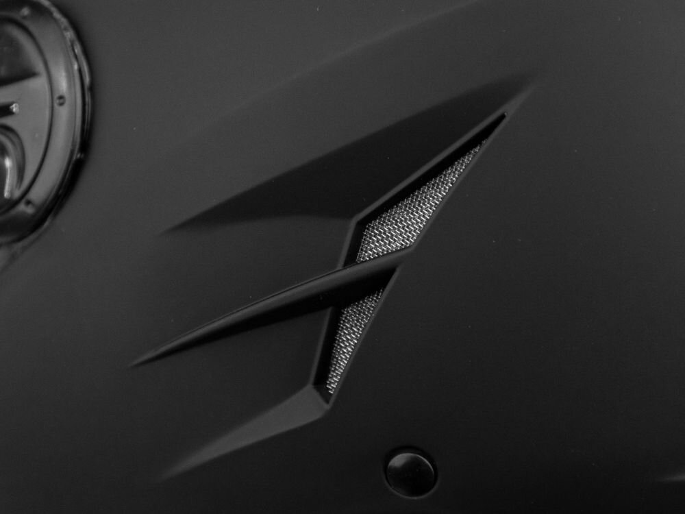 Zbliżenie na czarny kask motocyklowy z widocznym wlotem powietrza o geometrycznym kształcie i metalową siatką.