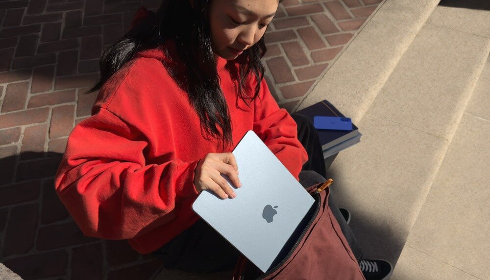 Osoba w czerwonej bluzie wkłada srebrny laptop z logo nadgryzionego jabłka do torby. W tle widać ceglaną nawierzchnię i kilka książek.