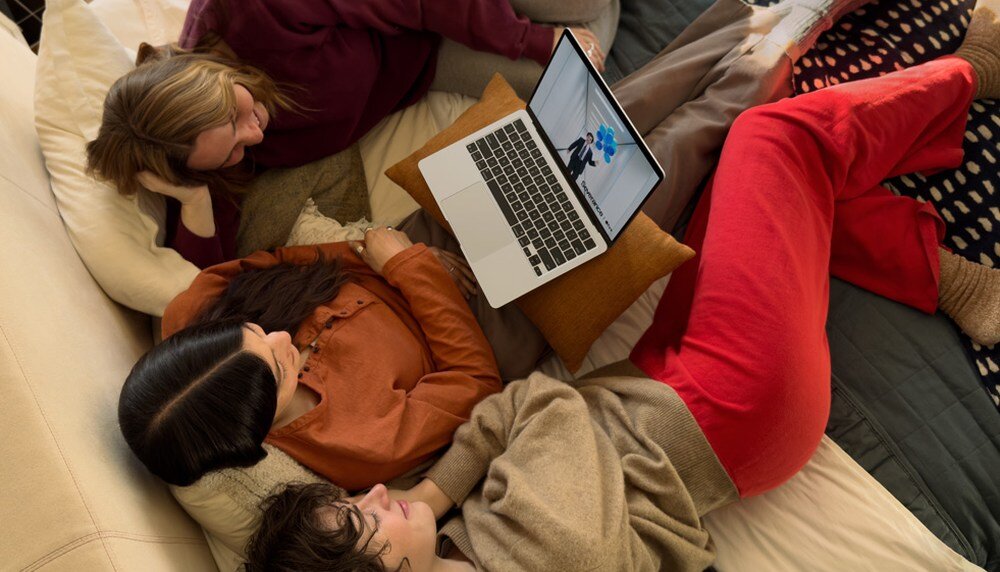 Trzy osoby leżą na łóżku, oglądając coś na laptopie. Laptop ma srebrną obudowę i wyświetla obraz na ekranie.