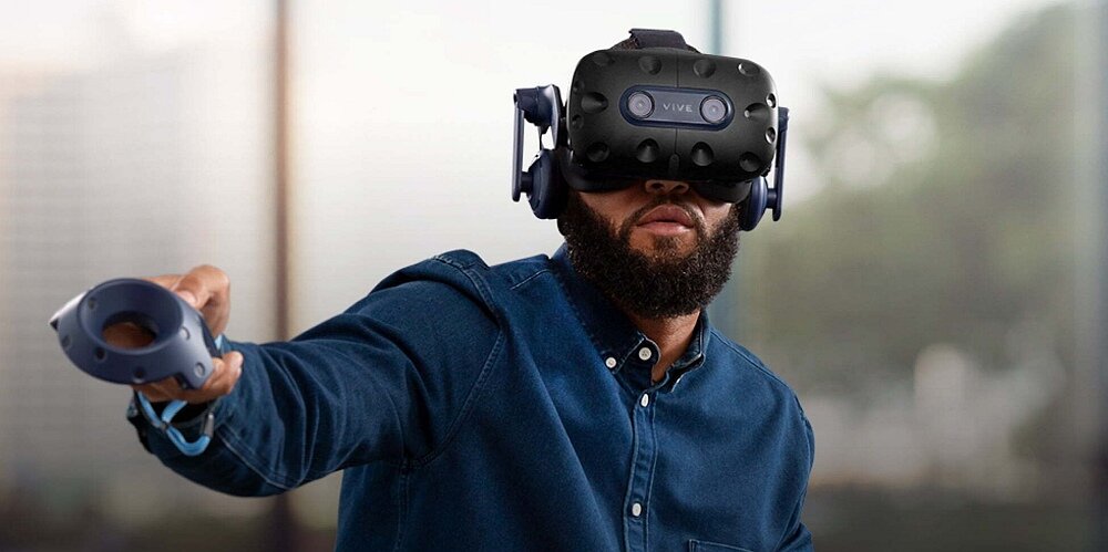 Osoba nosi gogle VR z napisem \'VIVE\' i trzyma kontroler w ręce. W tle widoczne jest rozmyte wnętrze pomieszczenia.