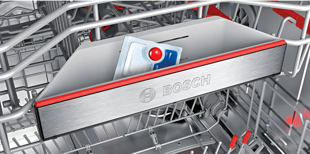 Wnętrze zmywarki z widocznym koszem na sztućce, w którym znajduje się tabletka do zmywania. Na froncie kosza widnieje logo \'BOSCH\'.