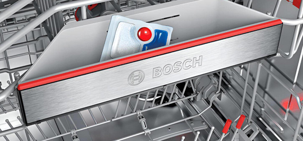 Wnętrze zmywarki z widocznym pojemnikiem na tabletki do mycia, na którym znajduje się logo BOSCH. W pojemniku umieszczona jest tabletka do zmywania z czerwonym elementem.