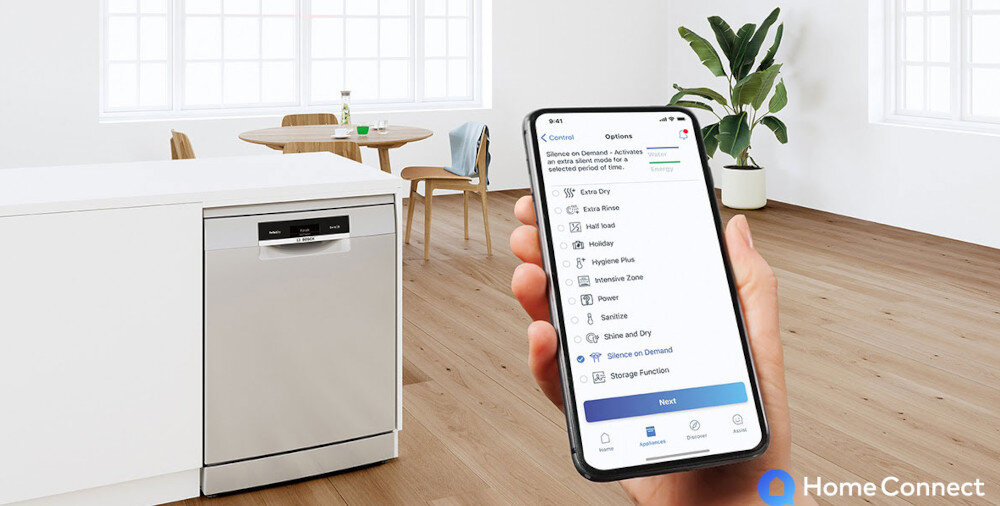 Na obrazie widoczna jest zmywarka w kuchni oraz ręka trzymająca smartfon z aplikacją Home Connect. W tle znajduje się stół z krzesłami i rośliną.