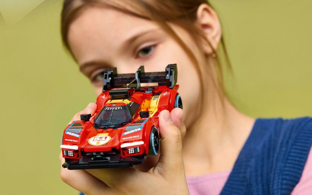 Osoba trzyma model samochodu wyścigowego z klocków LEGO w kolorze czerwonym z widocznym logo Ferrari na masce. W tle jednolite, zielone tło.