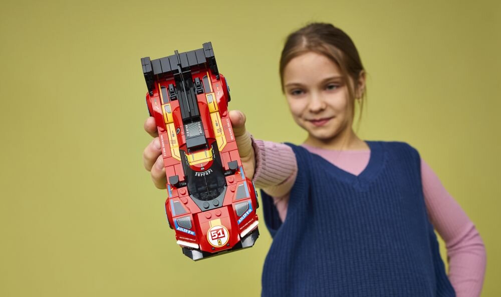 Osoba trzyma model samochodu z klocków LEGO w kolorze czerwonym z numerem 51 na przodzie. W tle jednolite, jasne tło.