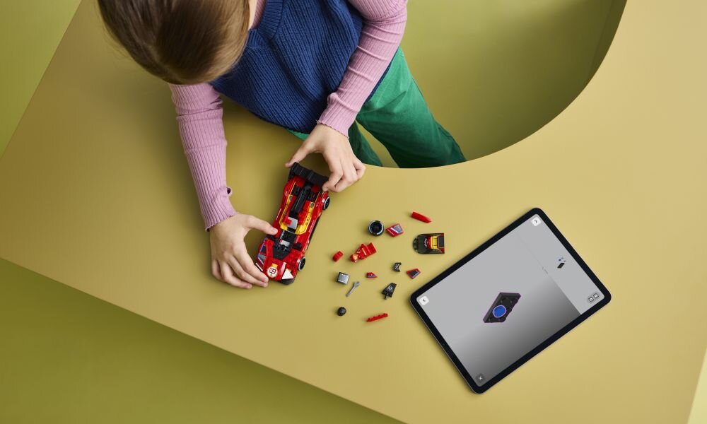 Dziecko bawi się czerwonym modelem samochodu z klocków LEGO na żółtym stole, obok leży tablet z wyświetlonym obrazem klocka LEGO.